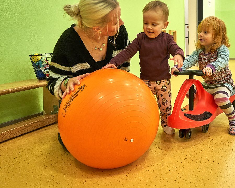 Eine Frau sitzt neben einem Kind auf einem Laufrad und ein Kind mit einem kleinen Gymnastik-Ball
